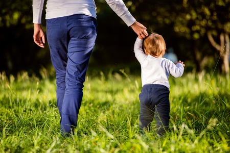 Unrecognizable mother holding hands of her little son making first steps outside in green sunny nature, back viewの写真素材