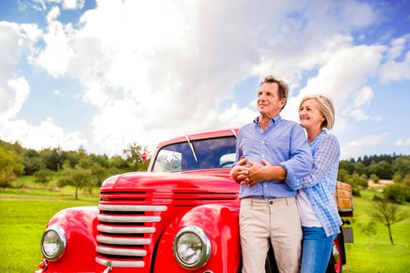 Senior couple hugging, vintage styled red car, green sunny natureの写真素材