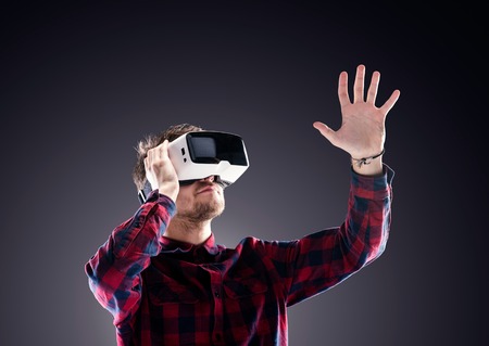 Hipster man in checked shirt wearing virtual reality goggles, reaching out. Studio shot on black...