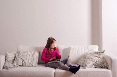 Little girl sitting on sofa with a laptop computer at home. Happy child playing indoors using PC, copy spaceの写真素材