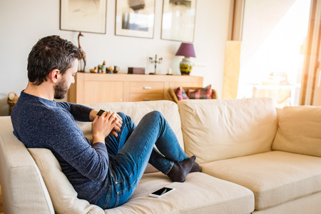 Man at home sitting on sofa using smart watch, smart phone laid on couchの写真素材