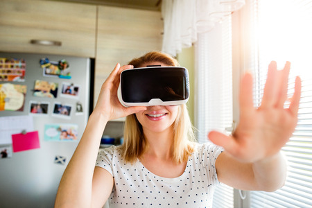 Blond woman wearing virtual reality goggles standing in a kitchen