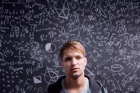 Handsome student in gray sweatshirt against big blackboard with mathematical symbols and formulas....