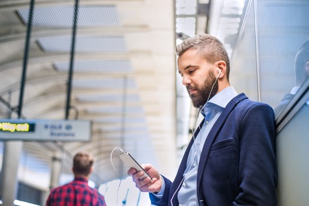Close up of handsome hipster businessman with smartphone standing on subway station listening musicの写真素材