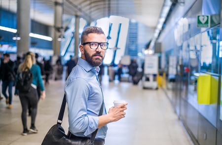 Handsome hipster businessman in blue shirt holding a coffee cup, standing on subway stationの写真素材