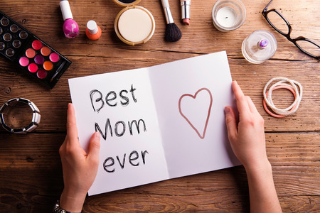 Hands of unrecognizable woman holding Mothers Day greeting card and various make up products. Studio shot on wooden background.の写真素材