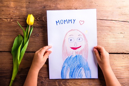 Mothers day composition. Hands of unrecognizable child holding a drawing of her mother and a yellow tulip. Studio shot on wooden background.の写真素材