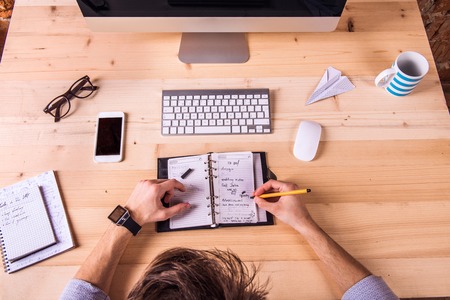 Businessman at the desk, wearing smart watch, writing into personal organizer.  Computer, smart phone and various office supplies around the workplace.の写真素材