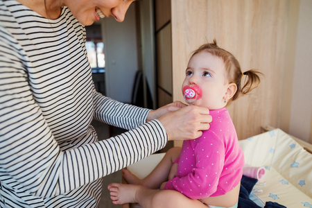 Beautiful young mother dressing her little daughter in the morning in her bedroomの写真素材