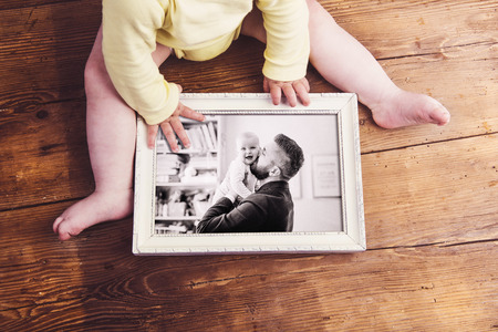 Fathers day composition. Unrecognizable baby holding a photo of father and daughter in white picture frame. Studio shot on wooden background.の写真素材