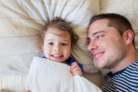 Young father having fun with his cute little daughter, lying in bedの写真素材
