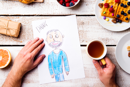 Fathers day composition. Hands of unrecognizable man with his childs drawing of him. Breakfast meal. Studio shot on wooden background.の写真素材