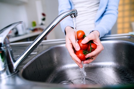 Hands of unrecognizable senior woman in checked blue shirt washing tomatoes under te tap. Preparing breakfast.の写真素材