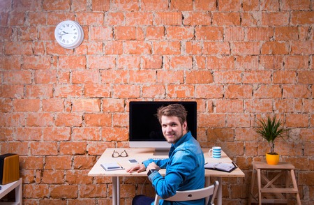 Hipster businessman sitting at office desk, smiling, against brick wall. Smart watch on hand and computer on the table. Coffee cup, personal organizer, smart phone and various office stuff around the workplace. Rear view.の写真素材