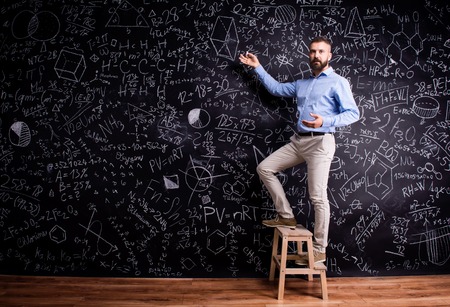 Hipster teacher writing on big blackboard with mathematical symbols and formulas, standing on step ladder. Studio shot on black background.の写真素材