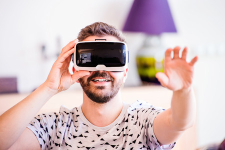 Young hipster man wearing virtual reality goggles, sitting in living room