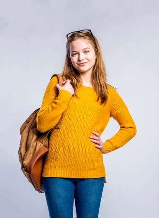 Teenage girl in jeans, yellow sweater, holding a backpack, young woman, studio shot on gray backgroundの写真素材