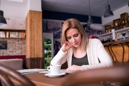 Blond woman with newspaper in cafe sitting at the table drinking coffee