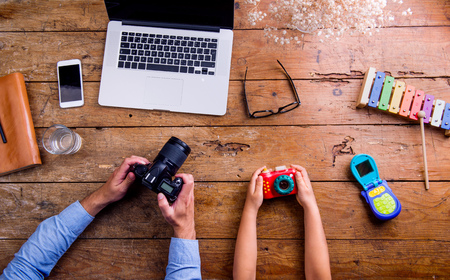 Hands of unrecognizable father and son holding cameras. Office desk with various office stuff around the workplace. Studio shot on wooden background. Flat lay.の写真素材