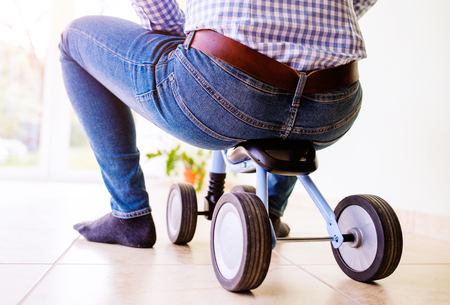 Unrecognizable hipster father in blue checked shirt and jeans riding a childrens bike indoors, sunny day, rear viewの写真素材