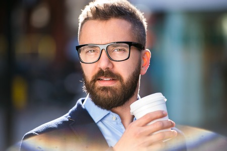 Hipster businessman holding a disposable cup and drinking coffee, walking in the street of London, wearing black eyeglassesの写真素材