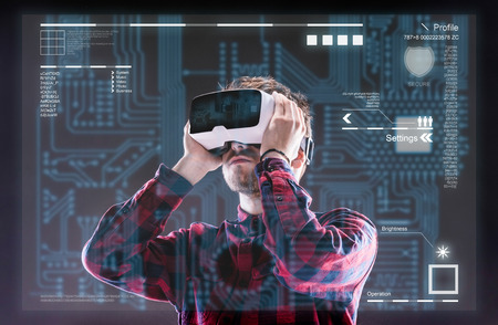Hipster man in checked shirt wearing virtual reality goggles, holding them. Studio shot on black...