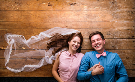 Beautiful young couple in love hugging, lying on a floor. Woman with veil, man with bow tie. Studio shot on wooden background. Copy space.の写真素材