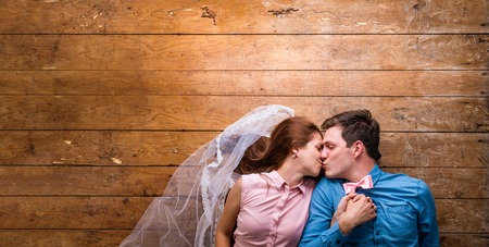 Beautiful young couple in love hugging, kissing, lying on a floor. Woman with veil, man with bow tie. Studio shot on wooden background. Copy space.の写真素材