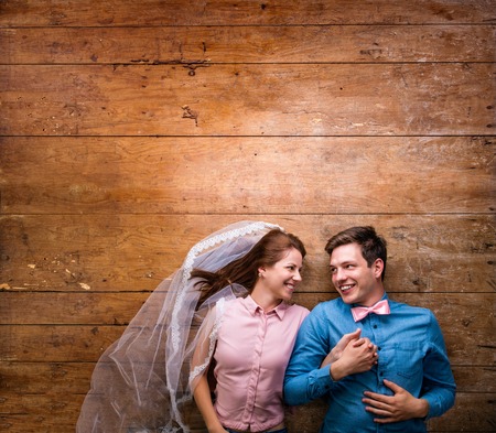 Beautiful young couple in love hugging, lying on a floor. Woman with veil, man with bow tie. Studio shot on wooden background. Copy space.の写真素材