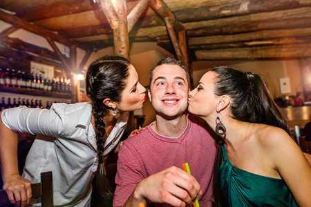 Two young beautiful women kissing man in bar or club having fun