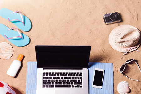 Summer vacation composition with laptop, smart phone, a pair of blue flip flop sandals, hat, sunglasses, sun cream and other stuff on a beach. Sand background, studio shot, flat lay, copy space.の写真素材