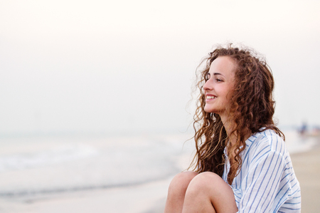 Beautiful young woman with curly hair sitting on beach wearing striped blue and white shirt, smiling. Enjoying time at seaside.の写真素材