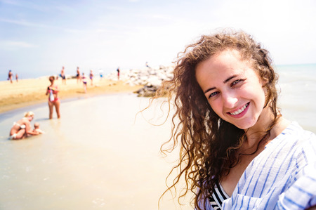 Beautiful young woman on beach wearing striped blue and white shirt, smiling, taking selfie. Enjoying time at seaside.の写真素材