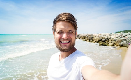Handsome hipster man in white t-shirt on beach, smiling, taking selfie. Enjoying time at seaside.の写真素材