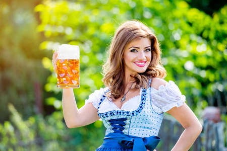 Beautiful young woman in traditional bavarian dress standing in the garden holding a mug of beer. Oktoberfest.の写真素材