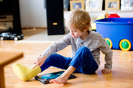 Cute little blond boy with broken leg in cast sitting on the wooden floor, playing on tablet....