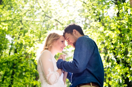Beautiful young wedding couple outside in green forest, touching foreheads. Bride in white dress and...