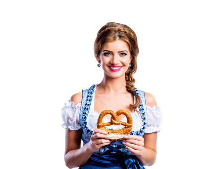 Beautiful young woman in traditional bavarian dress holding a pretzel. Oktoberfest. Studio shot on white background, isolated.の写真素材
