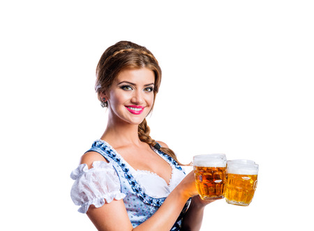 Beautiful young woman in traditional bavarian dress holding two mugs of beer. Oktoberfest. Studio shot on white background, isolated.の写真素材