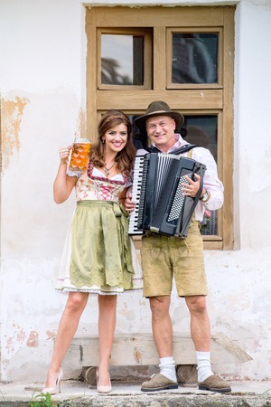 Couple in traditional bavarian clothes standing against old country house, man playing accordion, woman holding a mug of beer. Oktoberfest.の写真素材