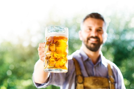 Handsome hipster young man in traditional bavarian clothes holding a mug of beer. Oktoberfest. Sunny summer garden.の写真素材