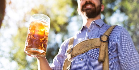 Unrecognizable young hipster man in traditional bavarian clothes holding a mug of beer. Oktoberfest. Sunny summer garden.の写真素材