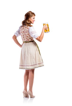 Beautiful young woman in traditional bavarian dress holding a mug of beer. Oktoberfest. Studio shot on white background, isolated. Rear viewの写真素材