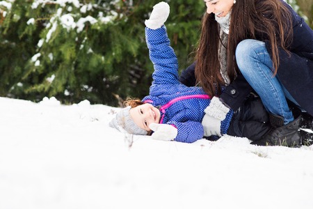 Beautiful young mother with her cute little daughter playing outside in winter nature, girl lying in the snowの写真素材