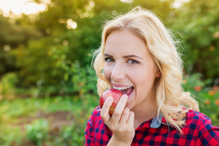 Beautiful young blond woman in red checked shirt eating apple. Autumn. Harvest.