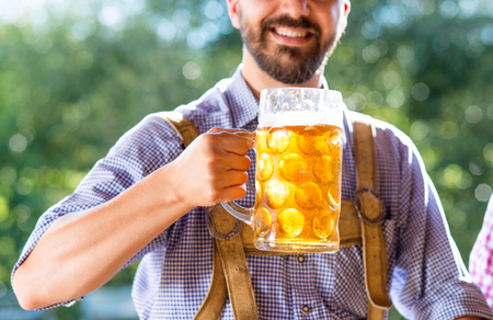 Unrecognizable young hipster man in traditional bavarian clothes holding a mug of beer. Oktoberfest. Sunny summer garden.の写真素材