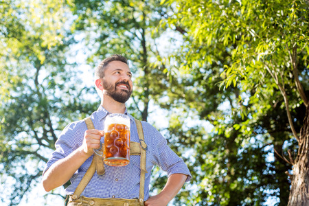 Handsome hipster young man in traditional bavarian clothes holding a mug of beer, arm on hip. Oktoberfest. Sunny summer garden.の写真素材