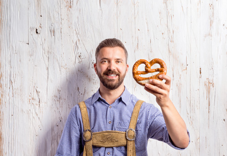 Handsome young man in traditional bavarian clothes holding pretzel. Oktoberfest. Studio shot on white wooden background.の写真素材