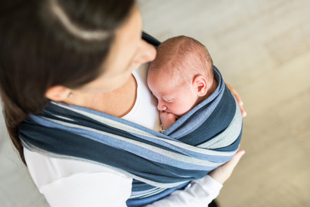 Close up of unrecognizable young mother with her newborn baby son in sling at homeの写真素材