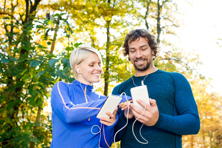 Beautiful couple running together outside in colorful sunny autumn forest using a fitness app on...